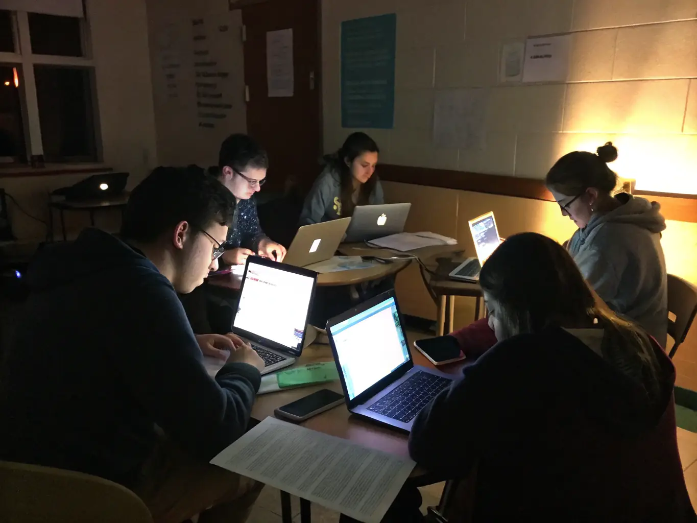 A diverse group of students collaborating on a machine learning project in a modern classroom setting, with laptops displaying complex code and data visualizations.